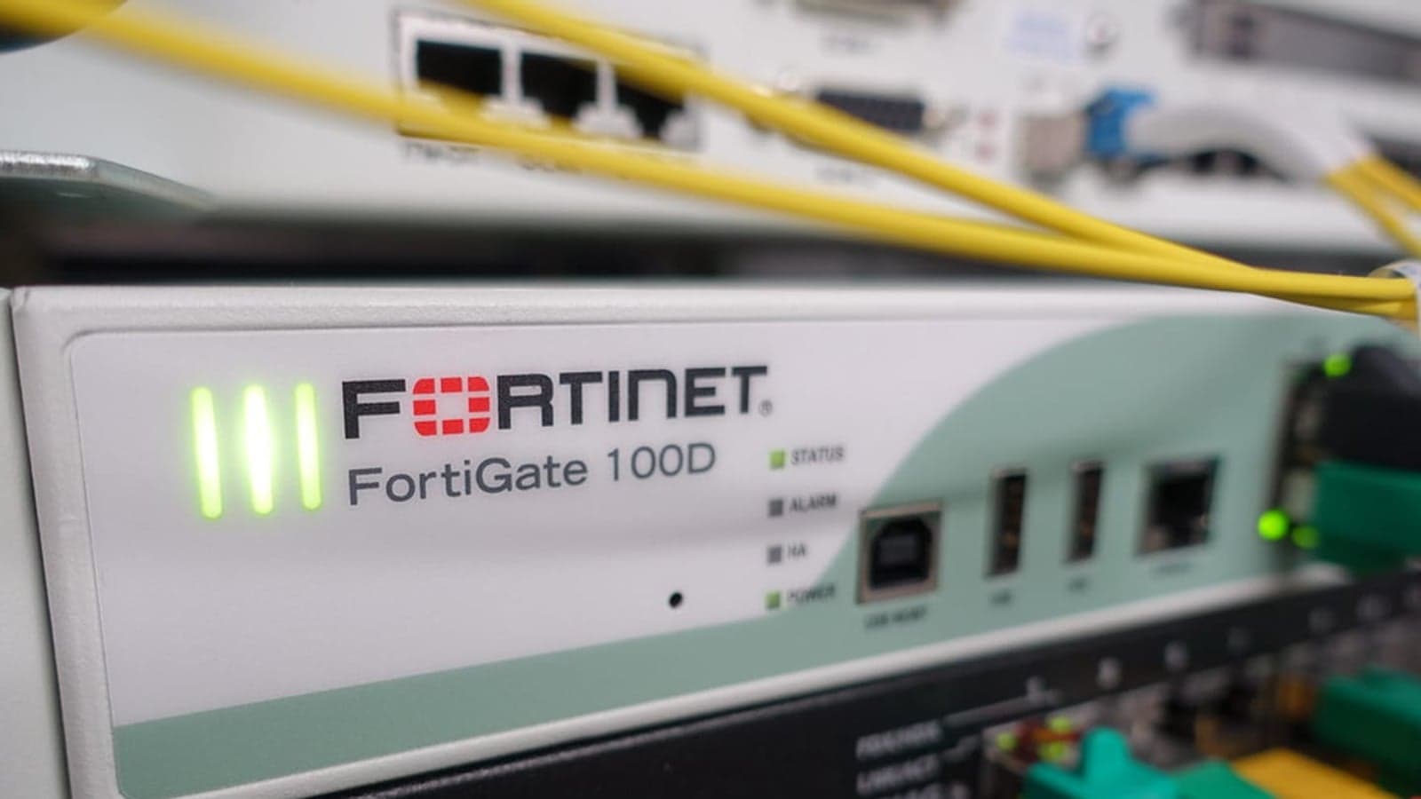 Frontal de un firewall Fortinet FortiGate 100D instalado en un armario de red, con sus luces verdes encendidas y cables de fibra amarillos por encima