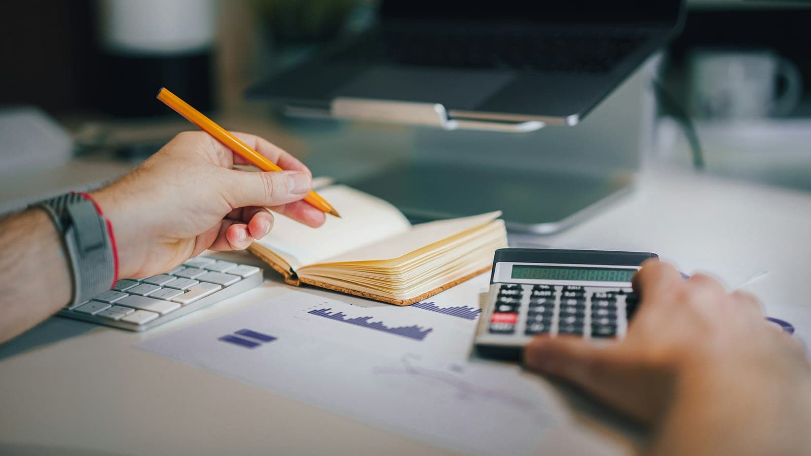 Hands with a pencil and a calculator going over accounts next to a notebook and a laptop